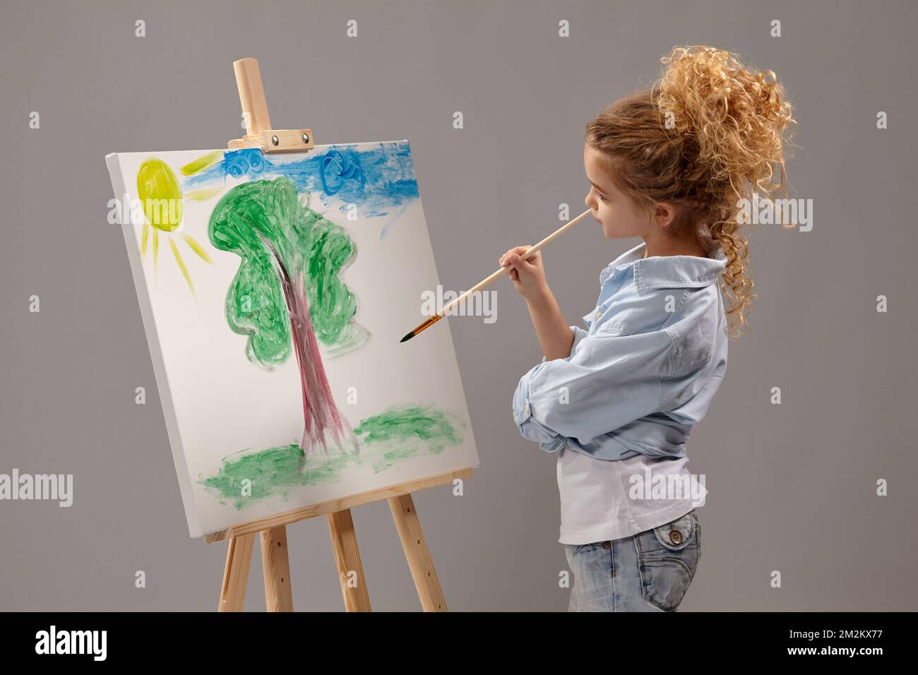 Charming school girl is painting with a watercolor brush on an easel, standing on a gray background. Stock Photo