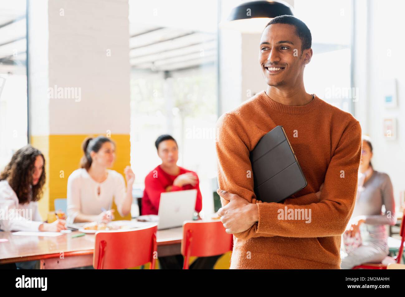 Attractive Moroccan young male in casual clothes standing and looking at his side smiling while holding a tablet. Teamwork sitting at desk on backgrou Stock Photo