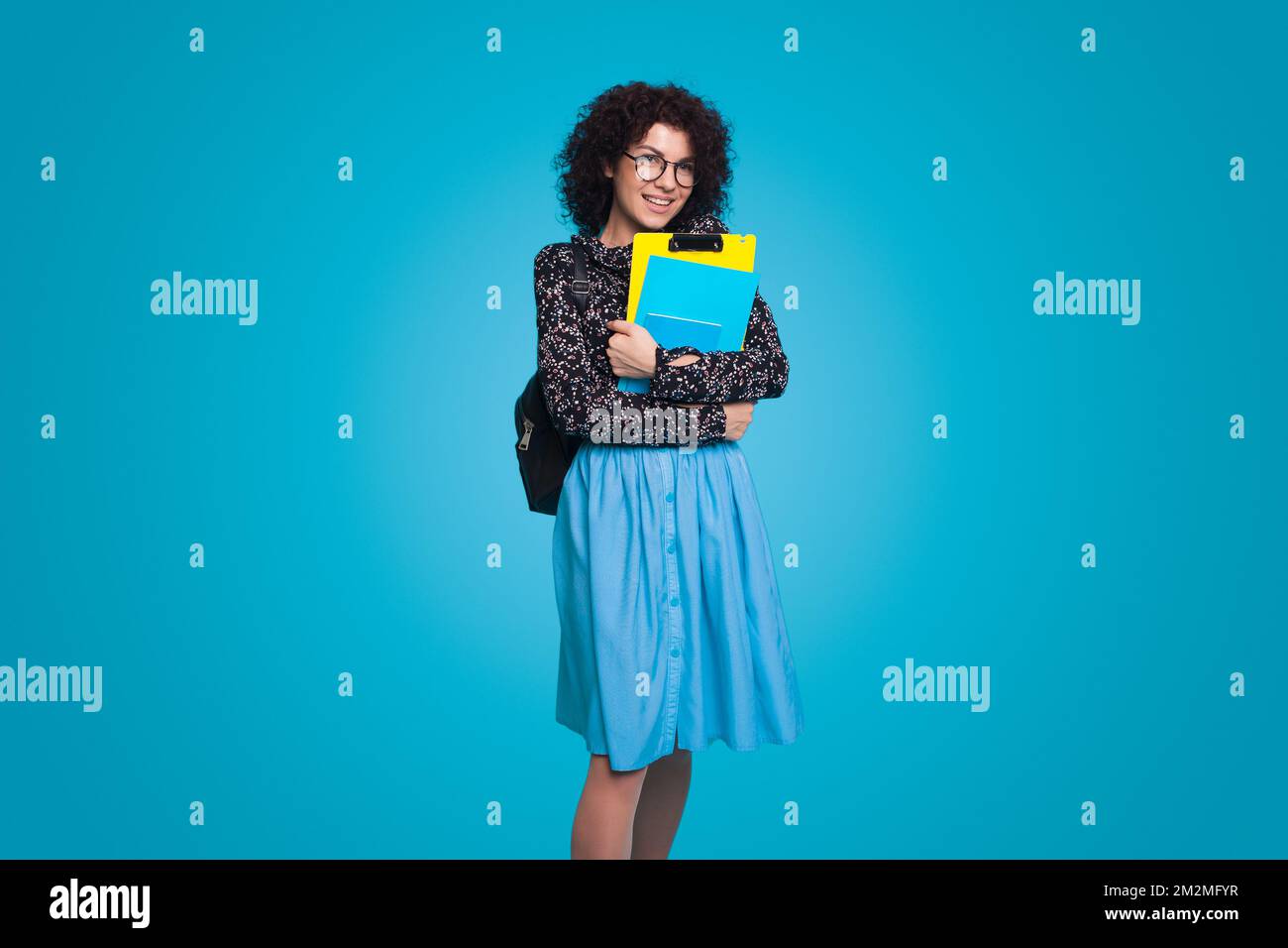 Young student woman with curly hair holding notebooks posing smiling isolated over blue background. . People lifestyle concept. Creative concept idea. Stock Photo