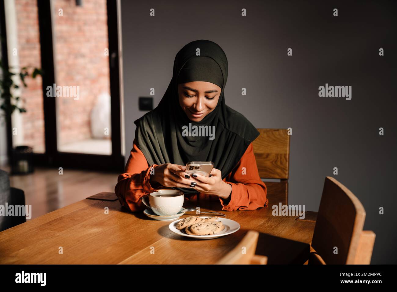 Young beautiful smiling woman in hijab with phone sitting at kitchen table with cup of tea and cookies Stock Photo