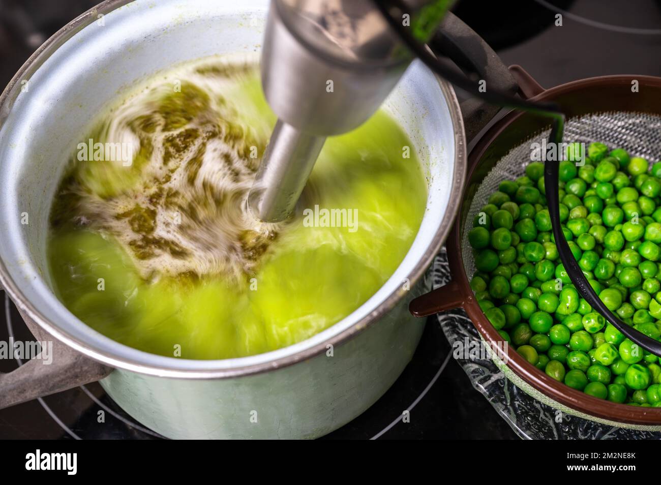 Stick blender blends pea soup in pot on hob, pea on strainer, closeup