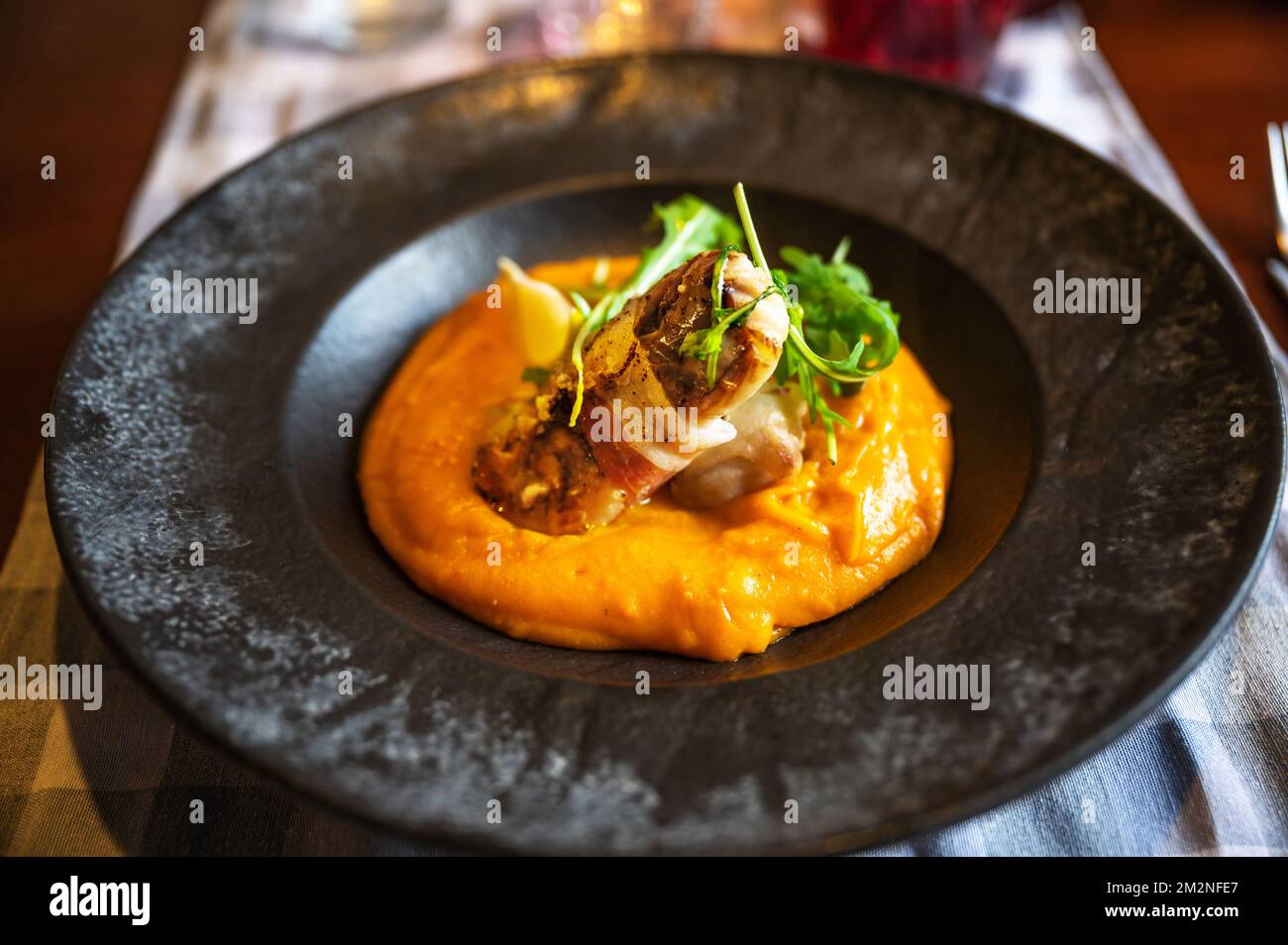Fried zander fillet with bacon on pumpkin puree, rucola leaf in black plate on restaurant table, closeup. Stock Photo