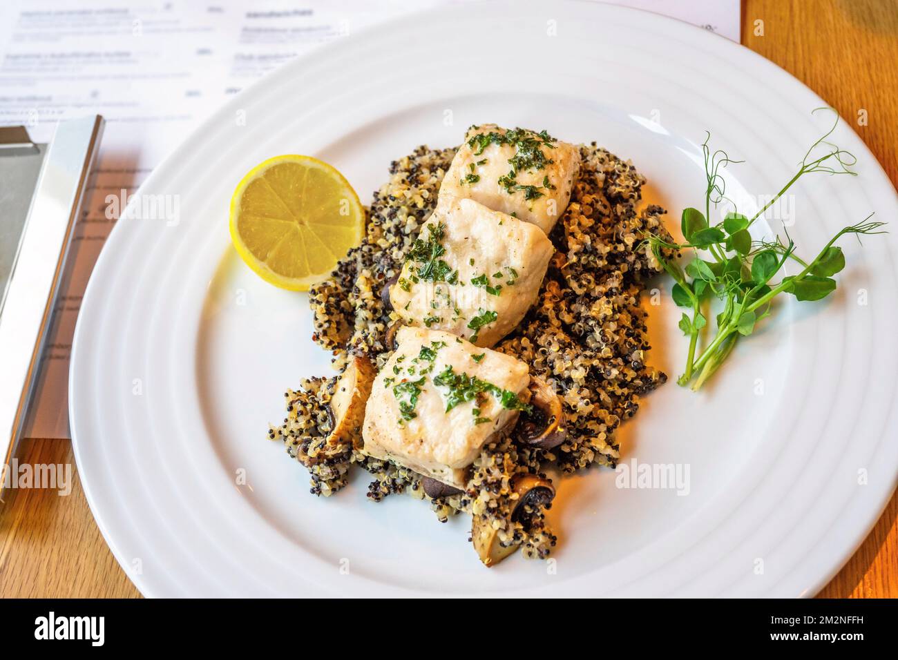 Baked sturgeon piece on quinoa with champignon mushroom, pea sprout and lemon on white plate, closeup. Dinner in restaurant. Stock Photo