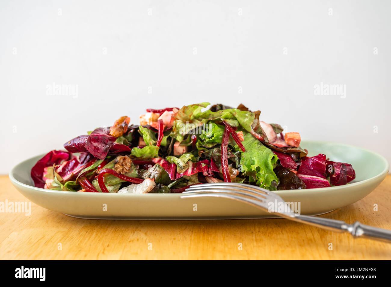 Red and green salad leaf with sliced beetroot, tofu cube and walnut on oval plate, fork on wooden table, closeup. Stock Photo