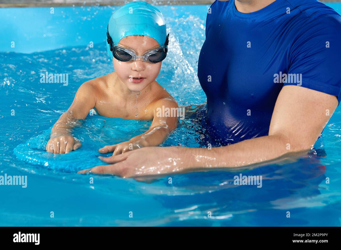 Little boy learning to swim in indoor pool with pool board and trainer Stock Photo