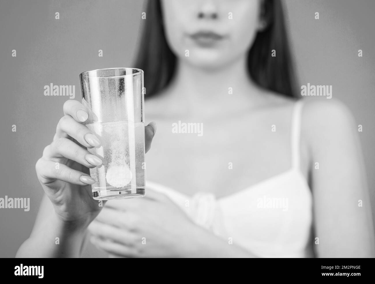 Woman taking drugs to releave headache. Brunette take some pills, holds glass of water, isolated on blue. Black and white Stock Photo