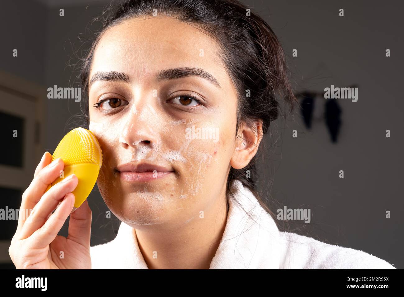 Millennial caucasian woman cleaning face skin with sponge and foam. Self skin care concept at home. Wearing bathrobe. Facial acne treatment idea. Stock Photo