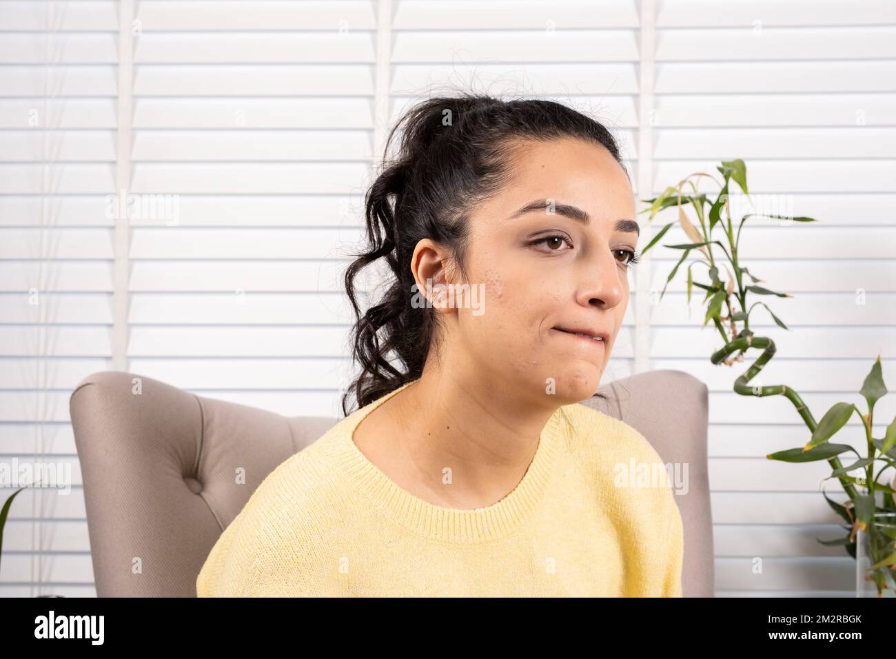 Sad woman, portrait of sad woman biting her lip. Pensive caucasian millennial female sitting on bergere in the living room. Pondering, thinking, lost. Stock Photo