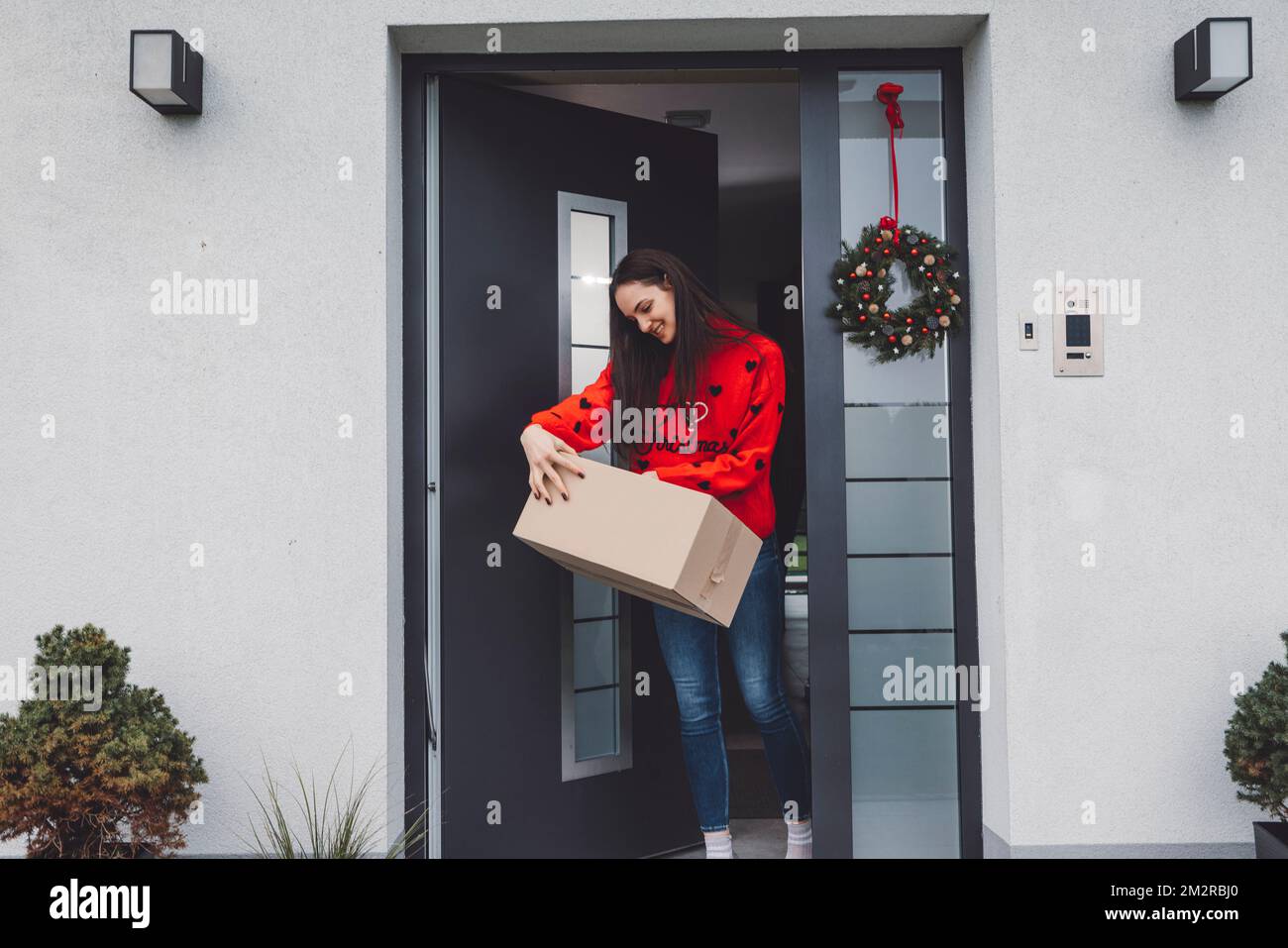 Happy young woman dressed in red jumper holding a delivery package on the front door taking it inside Stock Photo