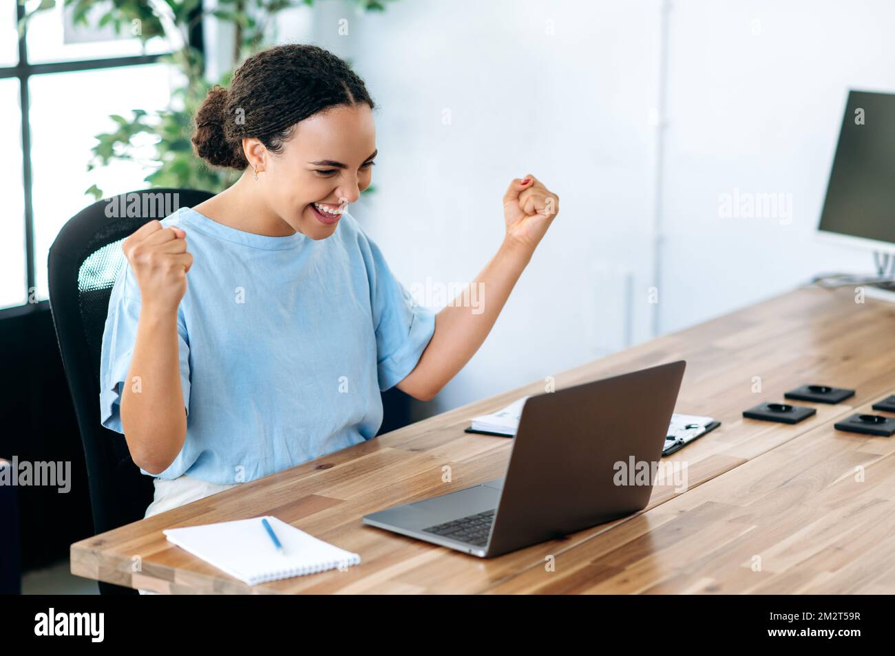 Photo of a joyful hispanic or brazilian business woman, sits at a work desk with laptop in modern office, rejoicing in success, big profit, celebrate deal, gesturing with fists, looks at laptop, smile Stock Photo