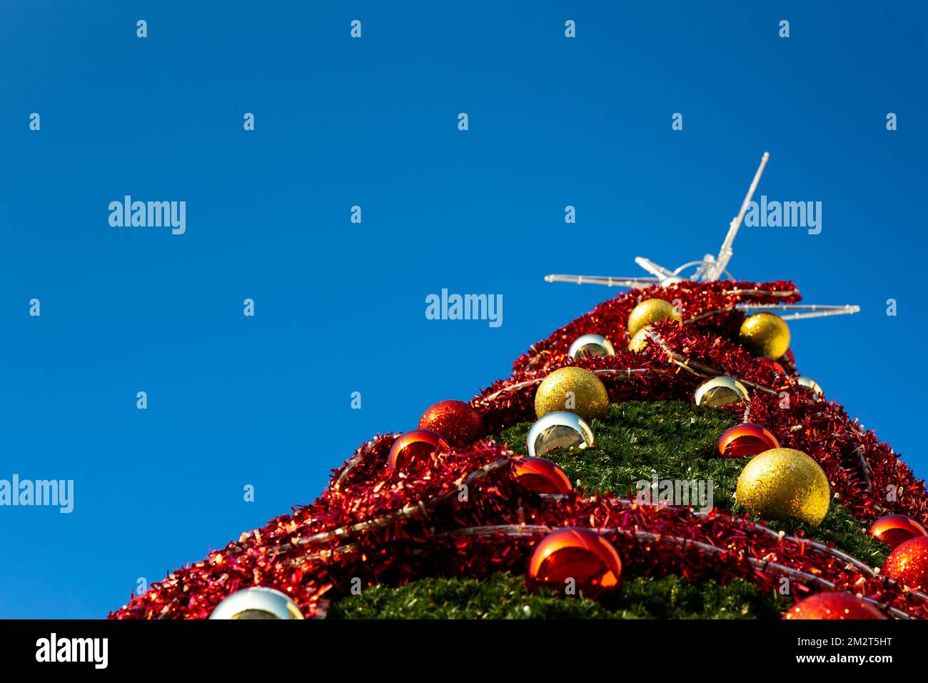 The top of an urban Christmas tree against a blue sky. Artificial green Christmas tree dressed in lights, red decorations and Christmas tree bubbles Stock Photo