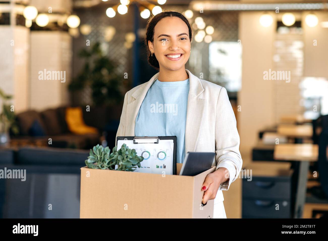 New job. Excited positive hispanic female employee, trainee, holding cardboard box, stand in modern office and looking at camera, smiling, newcomer woman have her first day of work in new workspace Stock Photo