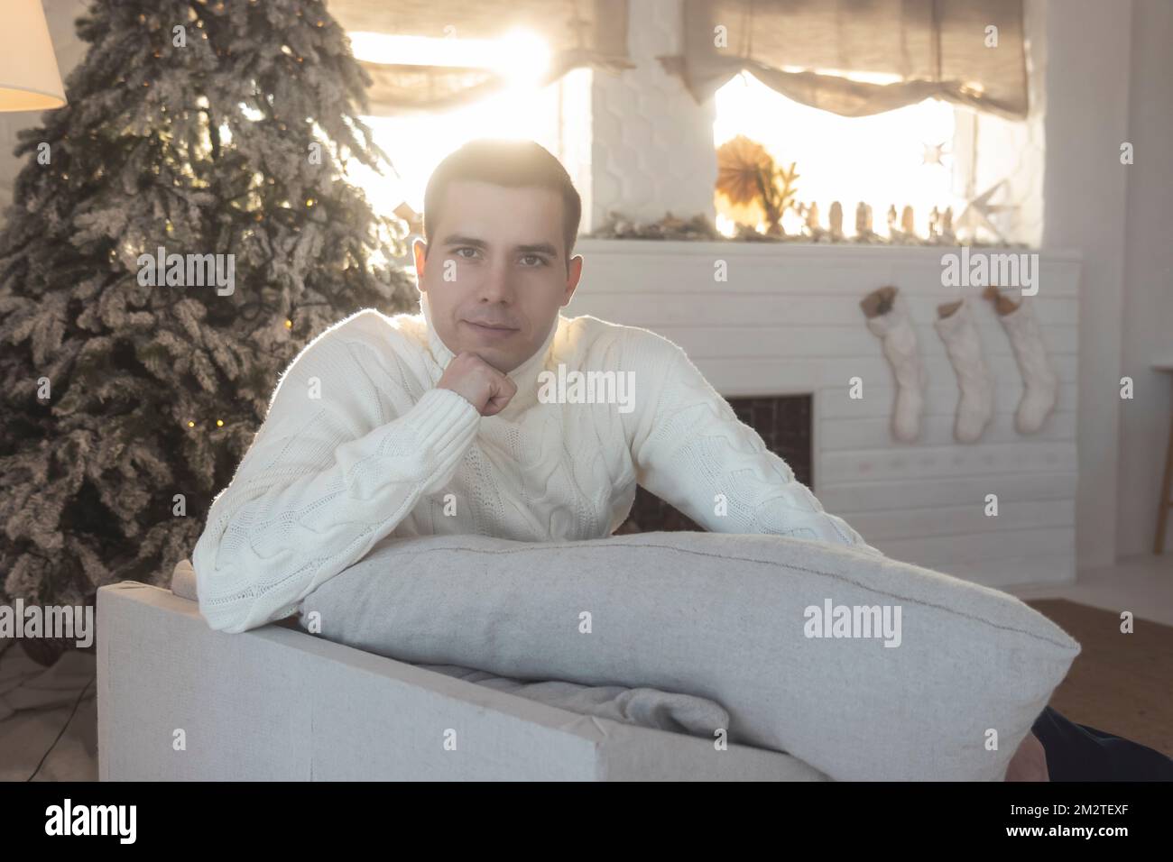 a man in a white sweater against the background of New Year's decorations sits in an armchair Stock Photo
