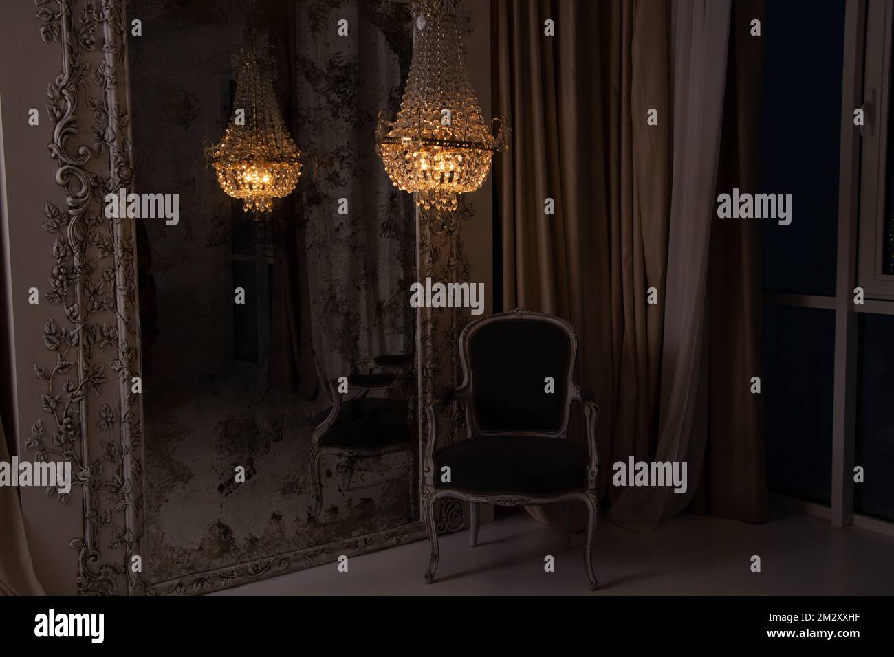 A crystal old chandelier hangs near the mirror and there is a chair next to the house in the evening near the window, apartment interior Stock Photo