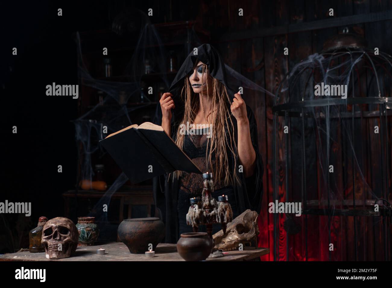 Witch dressed black hood with dreadlocks standing dark dungeon room use magic book for conjuring magic spell. Female necromancer occultist medium shoot Stock Photo