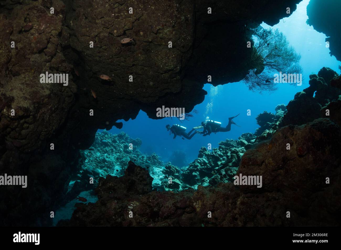 A view through the cave where two scuba divers swim along the reef. Stock Photo