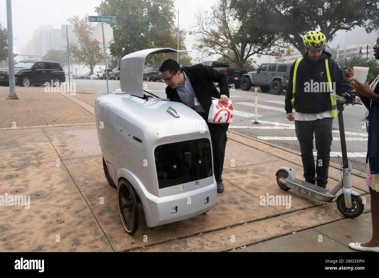 An autonomous delivery vehicle made by Austin-based Refraction AI delivers a Chick-fil-A lunch to  Travis County courthouse workers under the supervision of a test pilot that follows the vehicle on a scooter. The self-driving robots have been tested for months on Austin streets. Stock Photo