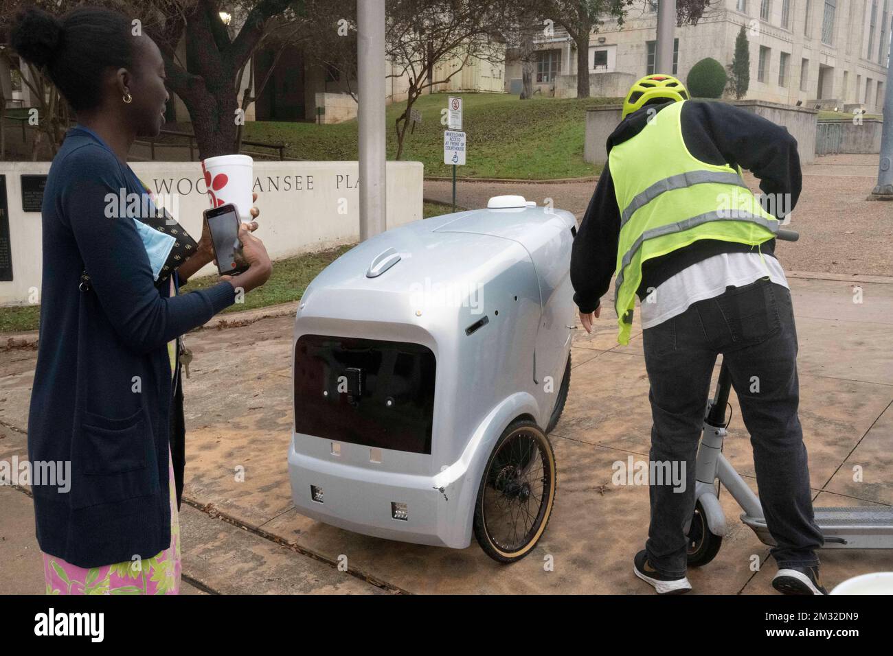 An autonomous delivery vehicle made by Austin-based Refraction AI delivers a Chick-fil-A lunch to  Travis County courthouse workers under the supervision of a test pilot that follows the vehicle on a scooter. The self-driving robots have been tested for months on Austin streets. Stock Photo