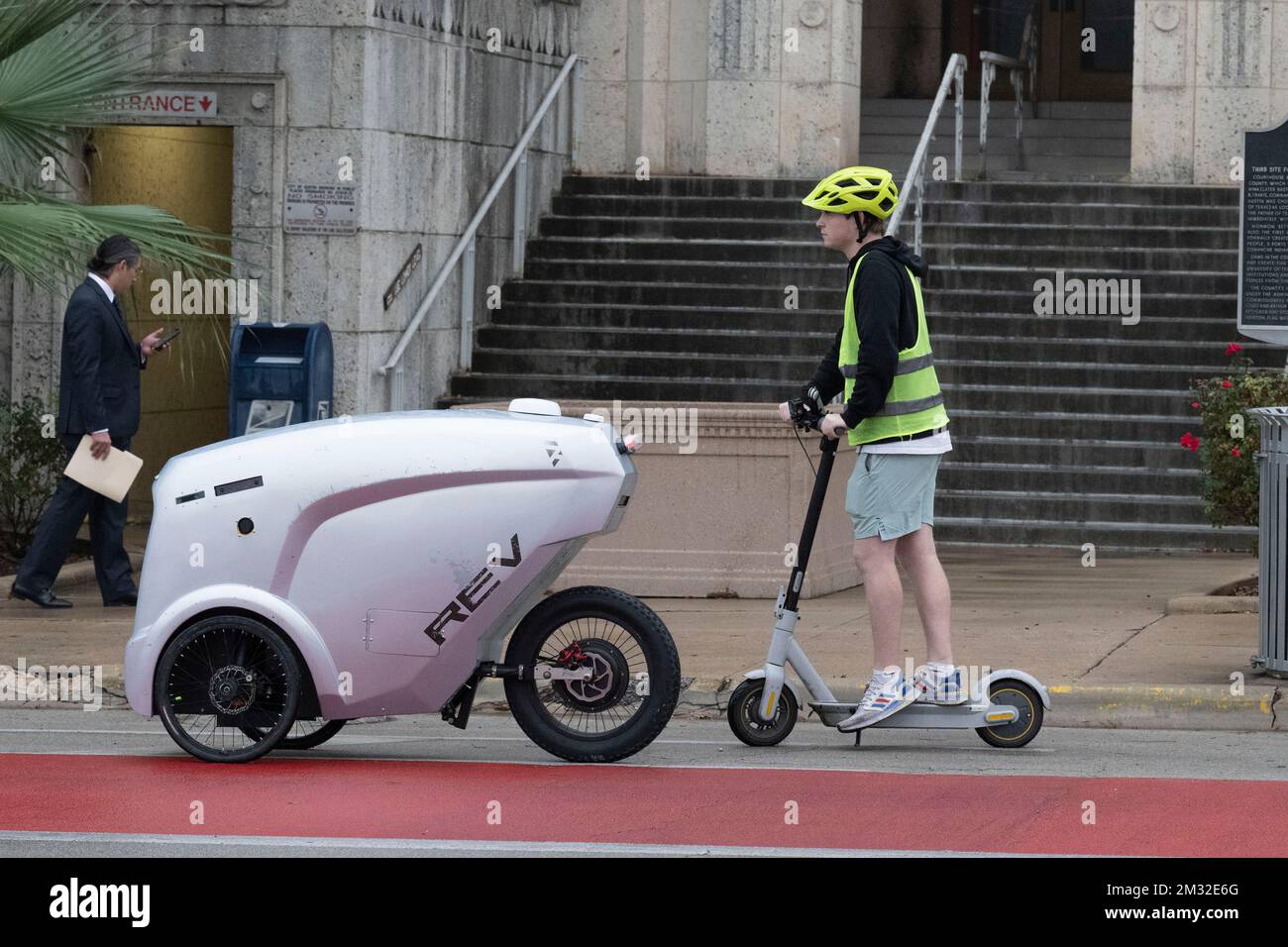 An autonomous delivery vehicle made by Austin-based Refraction AI delivers a Chick-fil-A lunch to  Travis County courthouse workers under the supervision of a test pilot that follows the vehicle on a scooter. The self-driving robots have been tested for months on Austin streets. Stock Photo