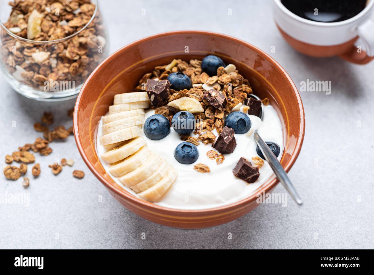 Yogurt with granola, banana, blueberries and dark chocolate in bowl, grey cocnrete table background, closeup view. Healthy food Stock Photo
