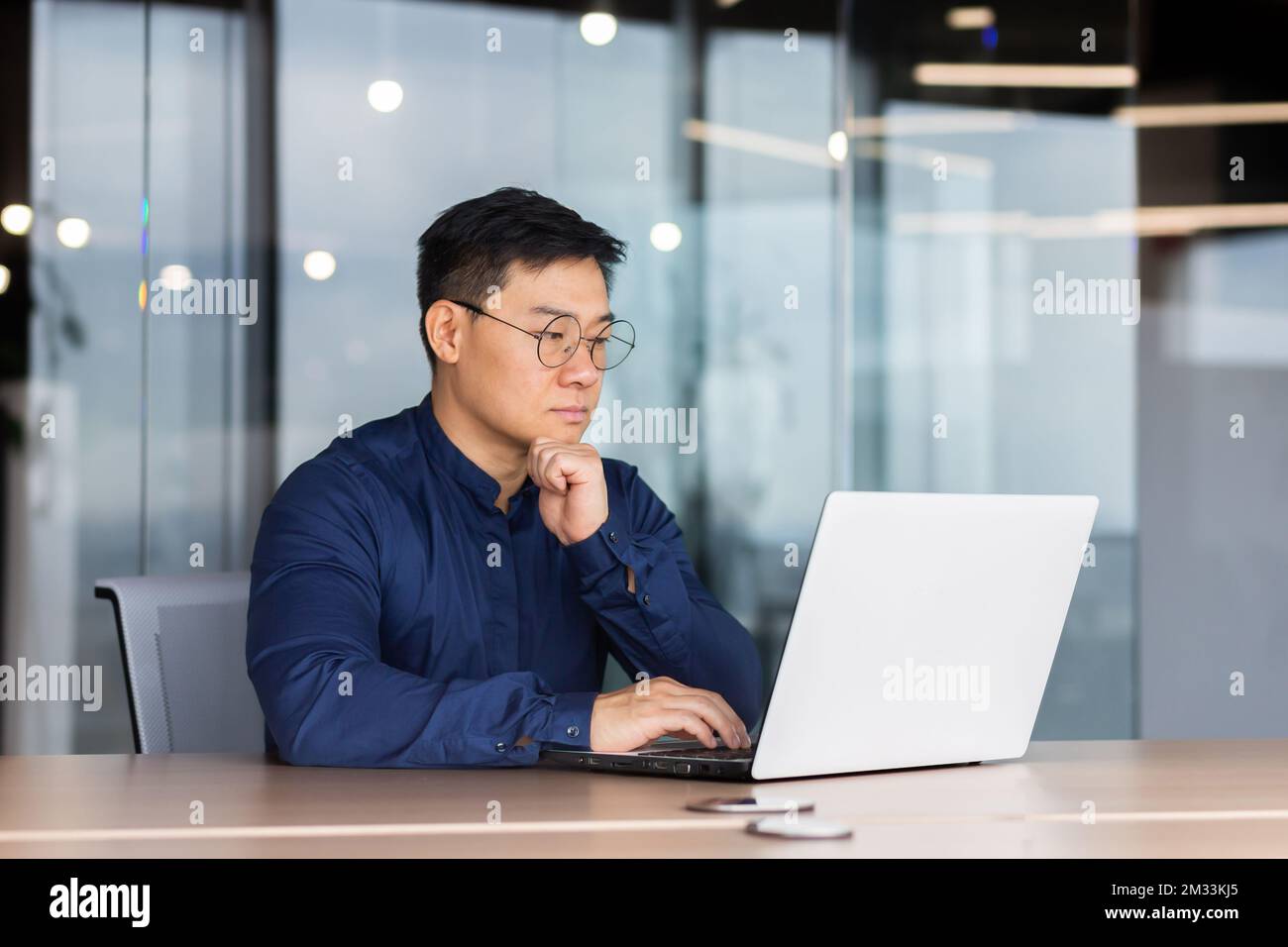 Serious and thoughtful businessman working inside office sitting at table using laptop at work, mature asian boss in shirt thinking and typing on keyboard. Stock Photo
