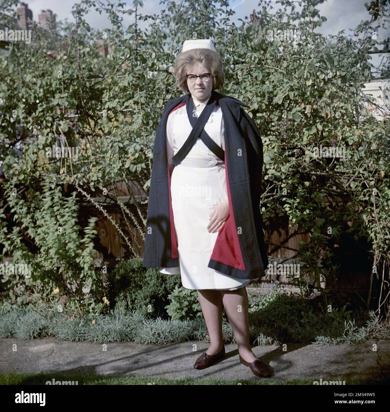 1960s, historical, standing outside for her photo, a young female nurse in her uniform of striped dress and white apron and over it, wearing a black cape, with cross strap fastening and maroon coloured lining, England, UK. The cape was designed to worn open at the front. On her head is a traditional nurse cap and she is wearing the spectacles that were fashionable in the 50s and 60s, known as Cat Eye glasses. Stock Photo