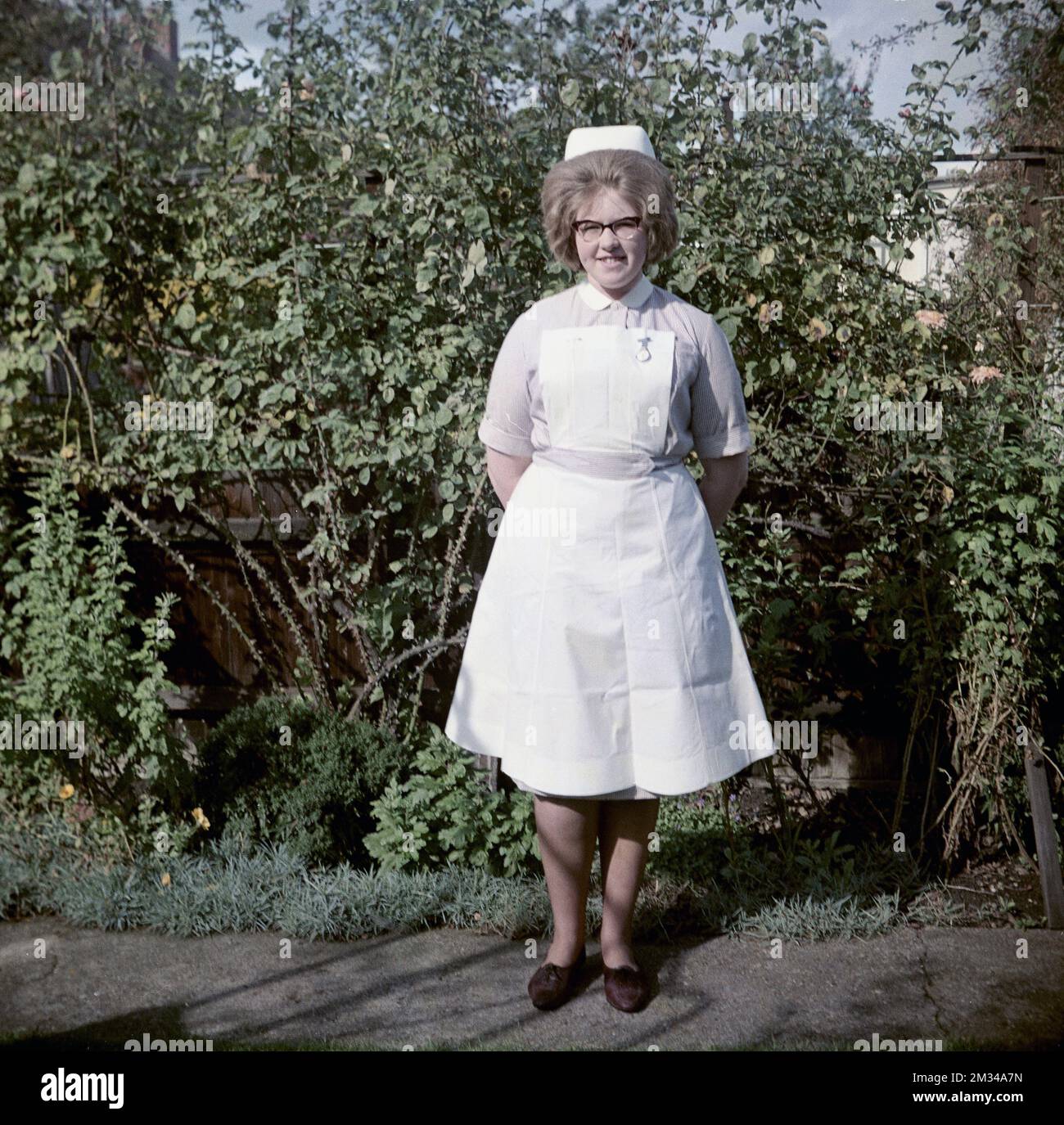 1960s, historical, standing outside for her photo, a young female nurse in her uniform, a  short sleeve striped lilac dress, with collar and white apron, with a nurses fob watch attached, England, UK. On her head is a traditional nurse cap and she is wearing the spectacles that were fashionable in the 50s and 60s, known as Cat Eye glasses. Stock Photo