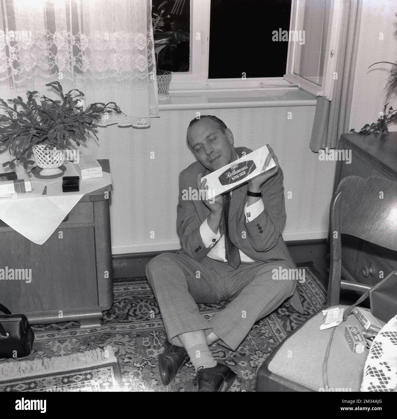 1960s, historical, a man in a suit & tie sitting on the floor of a front room of a house holding a carton of cigarettes, England, UK, acting like he is heaven! Stock Photo