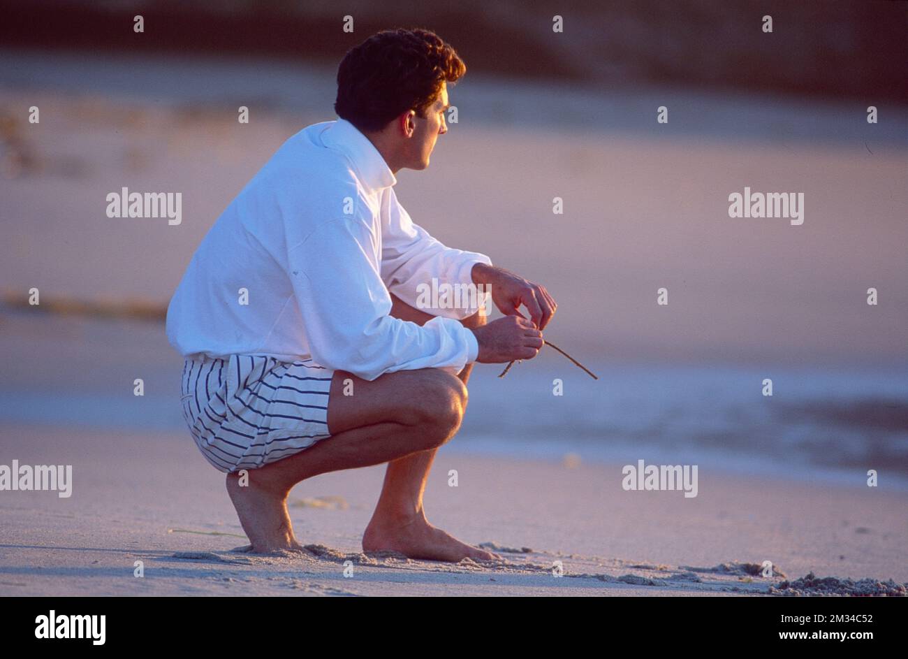 Man squatting down at the beach looking out over the water, pondering Stock Photo