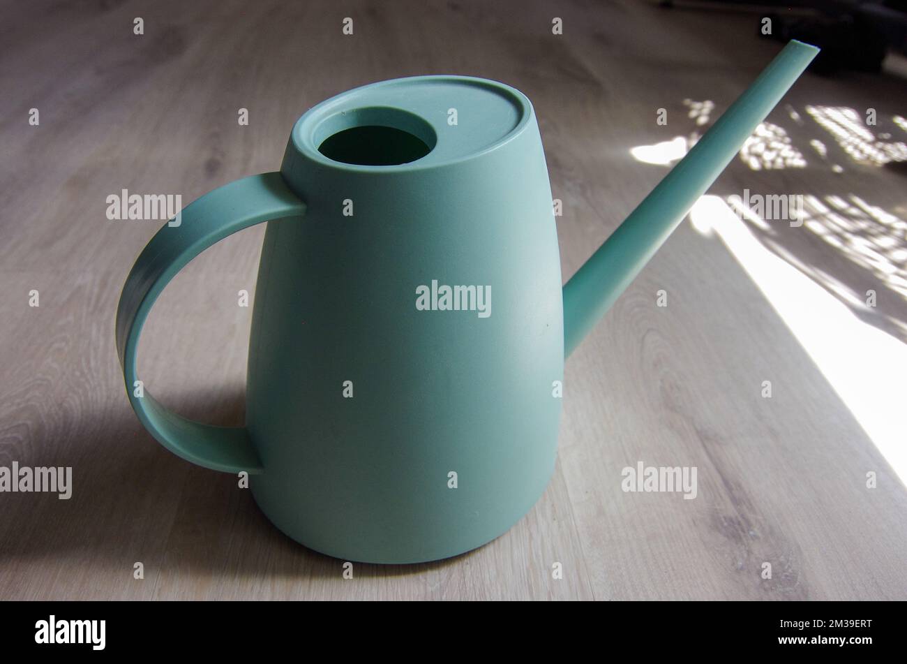 Turquoise blue watering can sitting on light wood floor with sunlight coming through the window Stock Photo