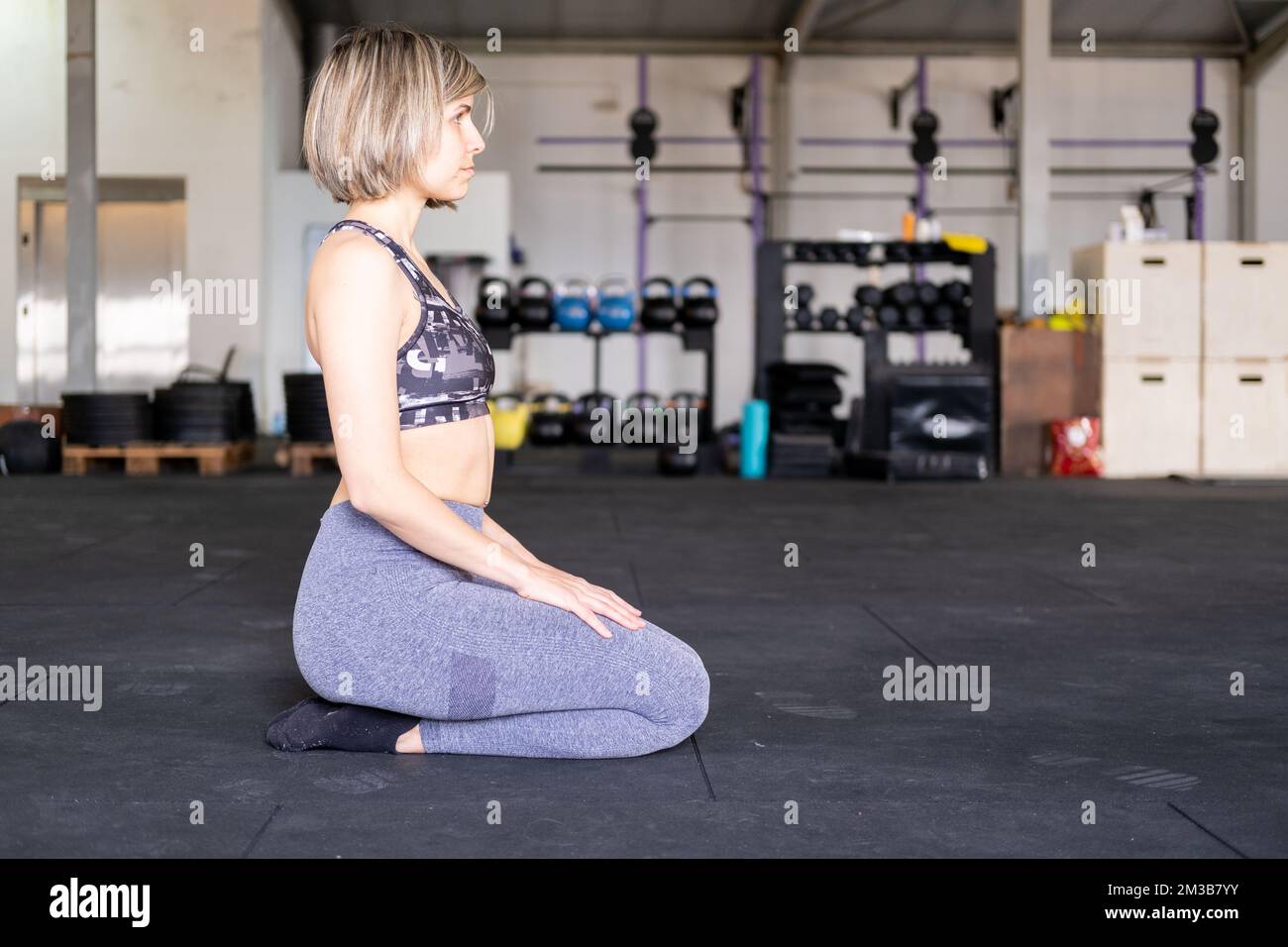 A blond mid adult woman resting in kneeling or vajrasana pose during her vinyasa flow yoga practice alone and wearing leggings and a sports bra in a g Stock Photo