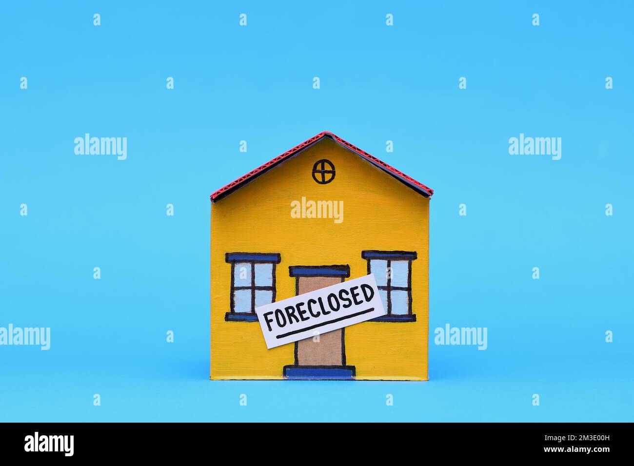 A yellow and red toy cardboard house with a Foreclosed sign on the door in the middle of frame isolated on a light blue background Stock Photo