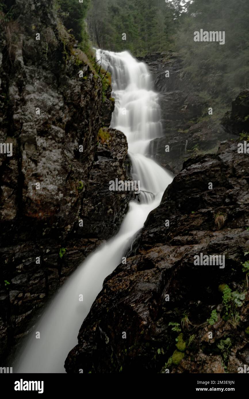 A long exposure of a waterfall flowing between rocky wet cliffs covered ...