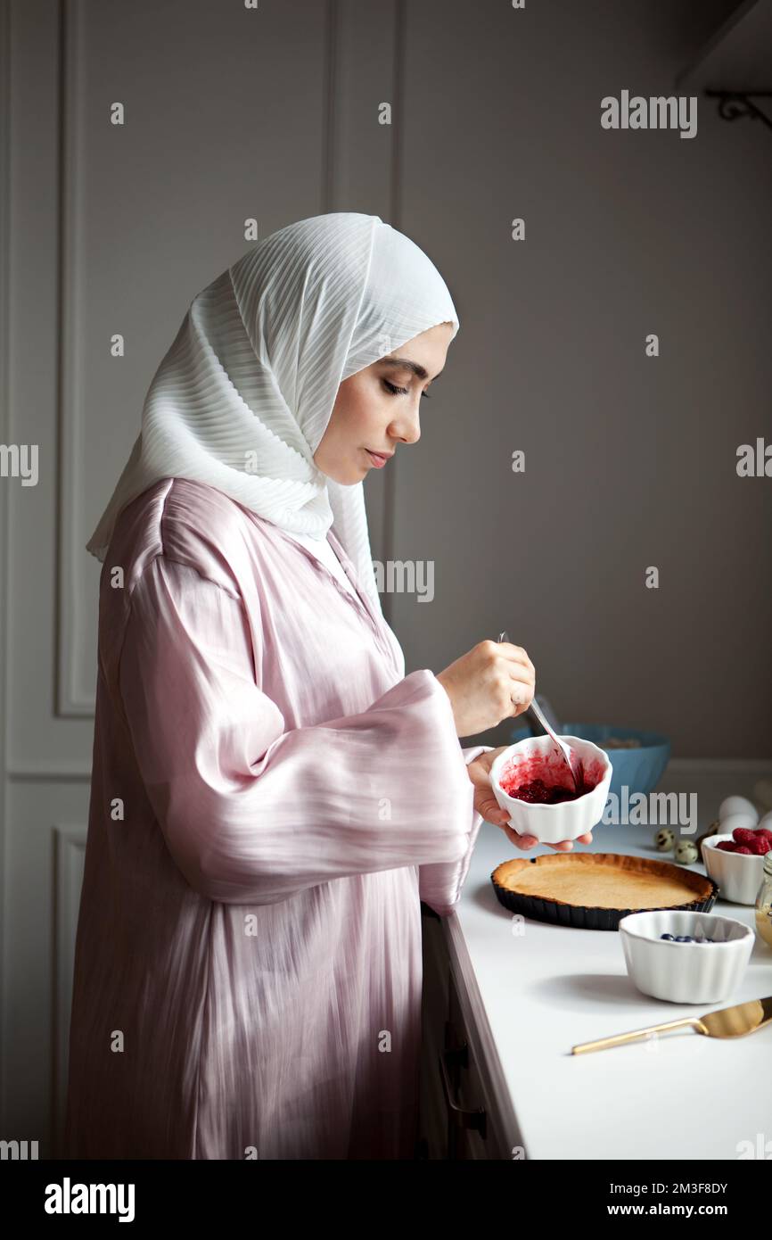 Side view portrait Muslim woman cooks dessert cake at kitchen, arabian young model in hijab and abaya. Islamic traditional clothing Stock Photo