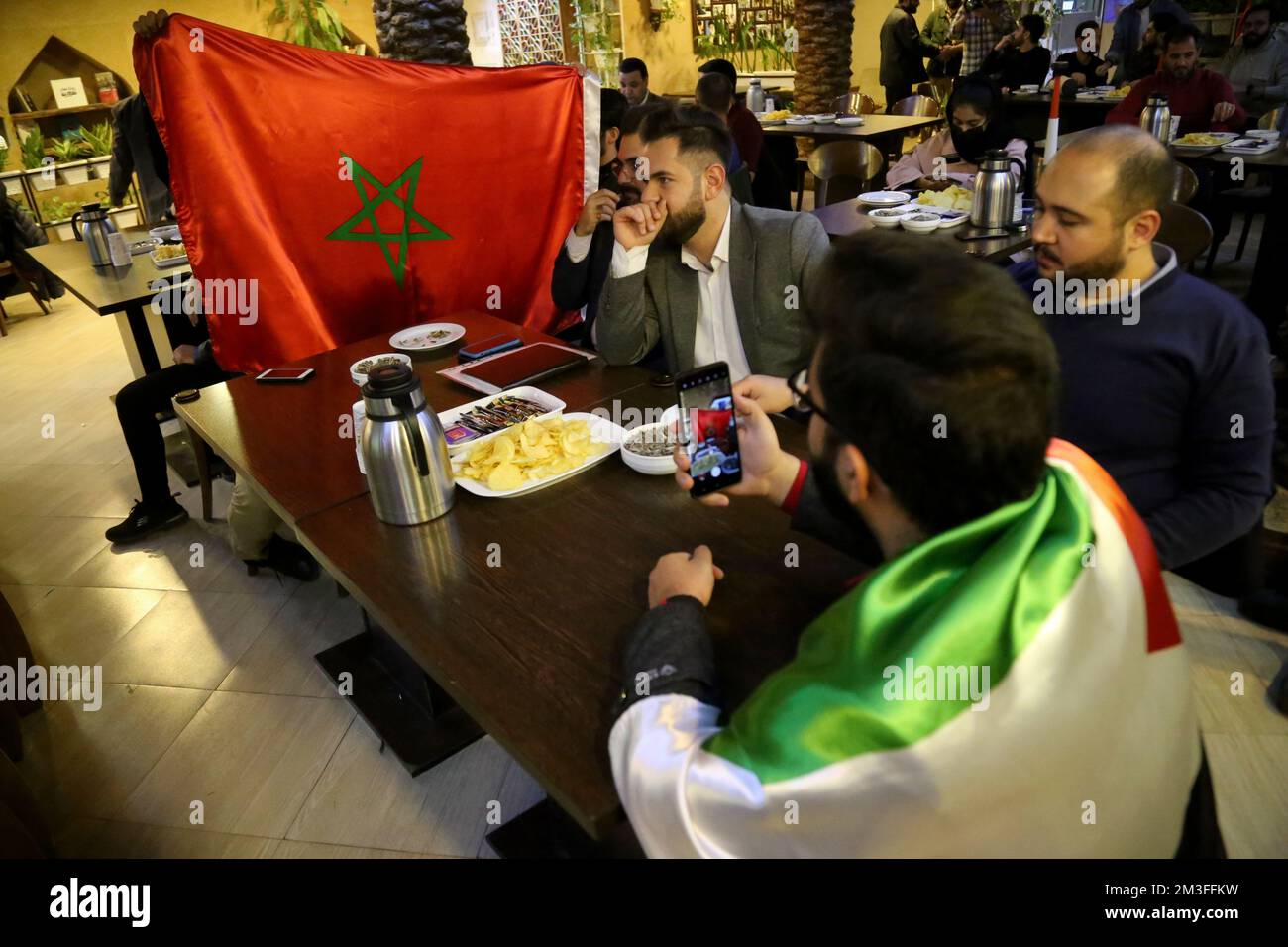 Tehran, Tehran, Iran. 14th Dec, 2022. An Iranian man holds a Moroccan flag while a group of Palestinian fans sitting behind them during the FIFA World Cup Qatar 2022 match Semi-Final between France and Morocco, at the Nakhlestan cafe in downtown Tehran, December 14, 2022. Fans from Iran, Lebanon, Yemen, Syria, and Palestine gather at the Nakhlestan cafe, which is run and owned by the Owj, Islamic Revolutionary Guard Corps (IRGS) Arts and Media Organization, to support the Moroccan national team. Following the win against Portugal in Qatar on December 10th, Moroccans carried Palestine flags Stock Photo