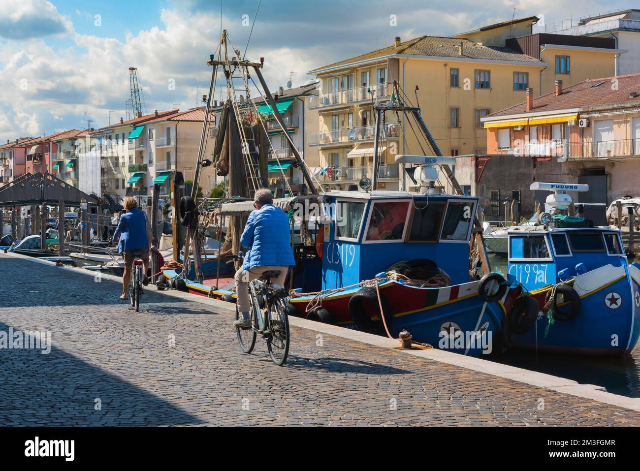 Senior couple cycling, rear view of two senior cyclists riding their bikes along a quay in an Italian fishing port, Chioggia, Veneto, Italy Stock Photo