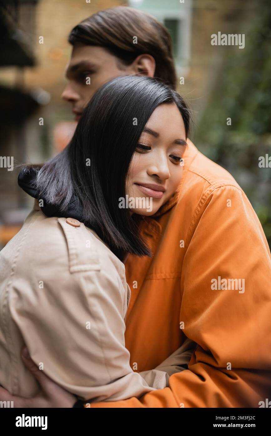 Young asian woman in trench coat hugging blurred boyfriend on terrace ...