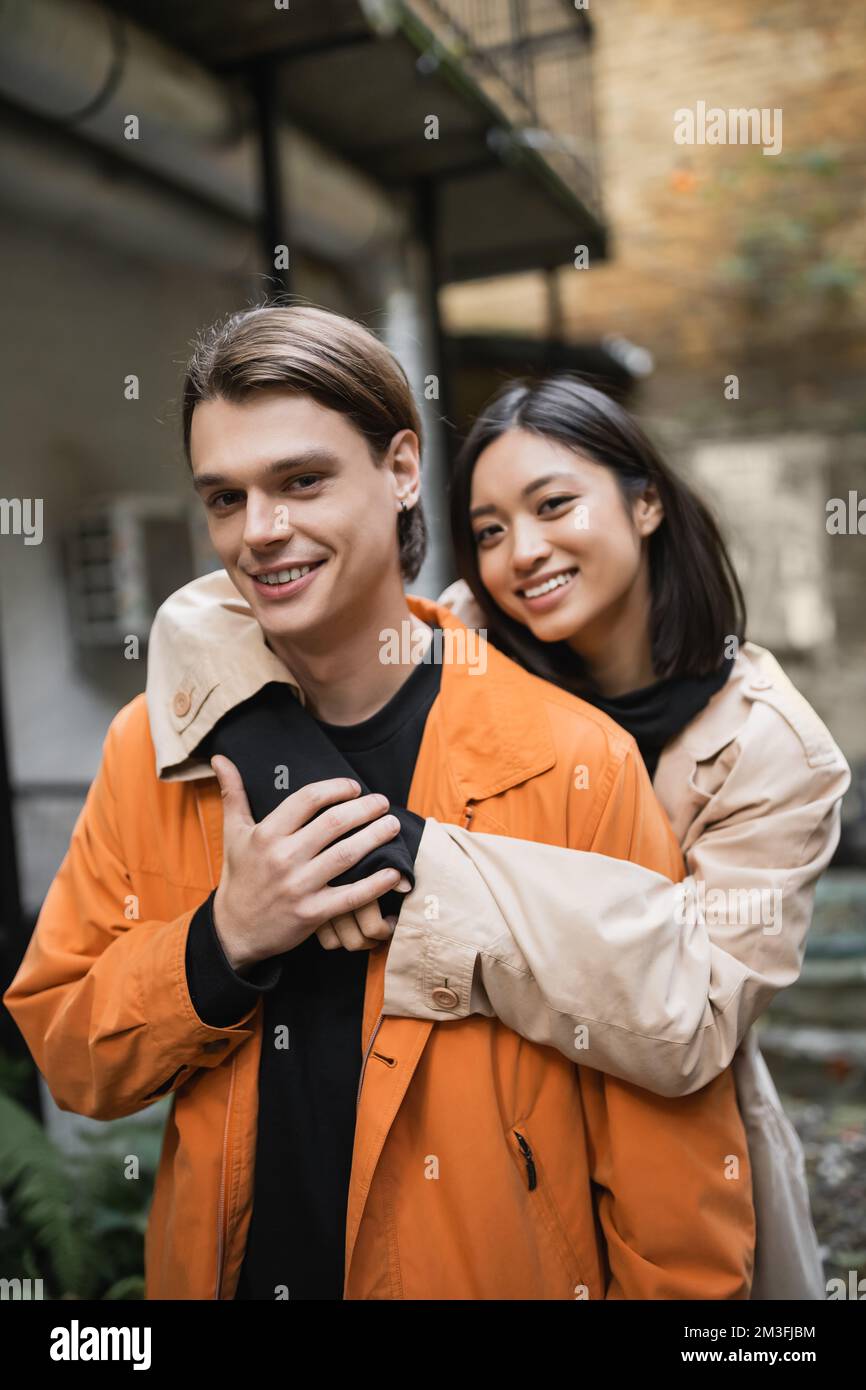 Cheerful asian woman hugging boyfriend and looking at camera in outdoor ...