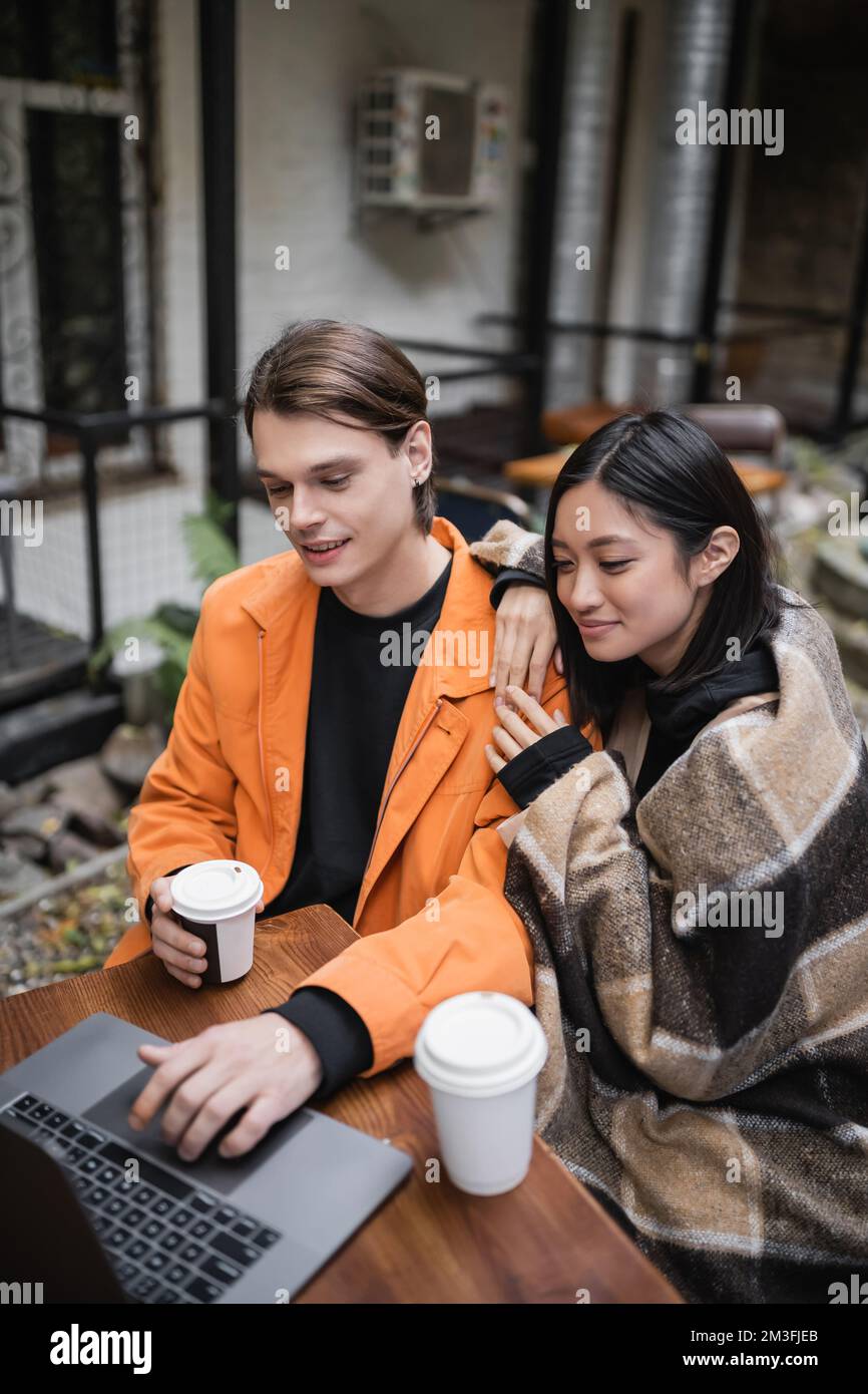 Positive asian woman in blanket hugging boyfriend using laptop near ...