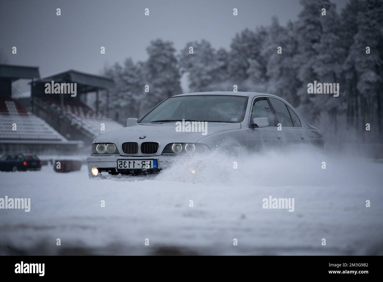 12-12-2022 Riga, Latvia  a car driving through a snow covered parking lot in the snow with trees in the background and a snow bank in the foreground. Stock Photo