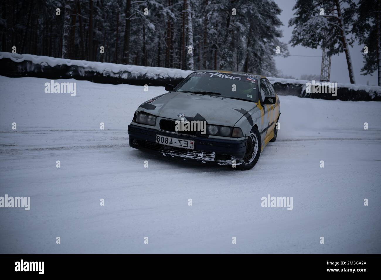 12-12-2022 Riga, Latvia  a car is driving through the snow in the woods on a road with trees in the background and a yellow and black stripe on the fr Stock Photo