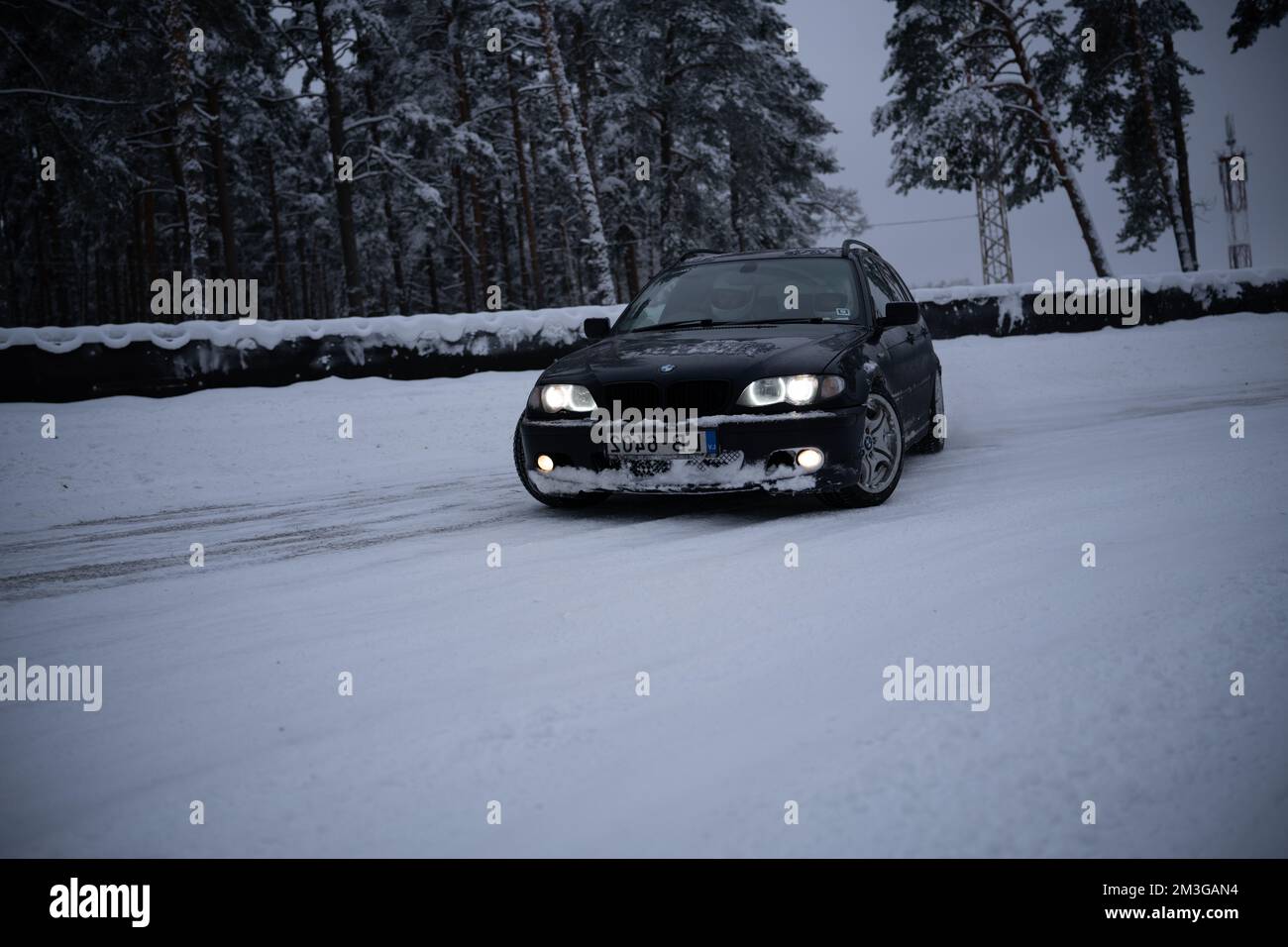 12-12-2022 Riga, Latvia  a car driving down a snowy road in the snow with headlights on and trees in the background in the snow. . Stock Photo