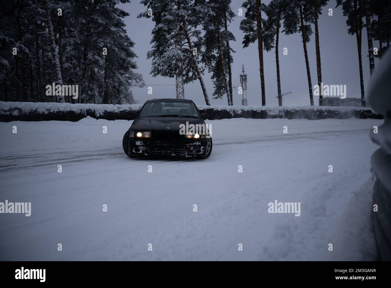 12-12-2022 Riga, Latvia  a car driving down a snowy road in the snow with headlights on and trees in the background and a person standing in the snow. Stock Photo
