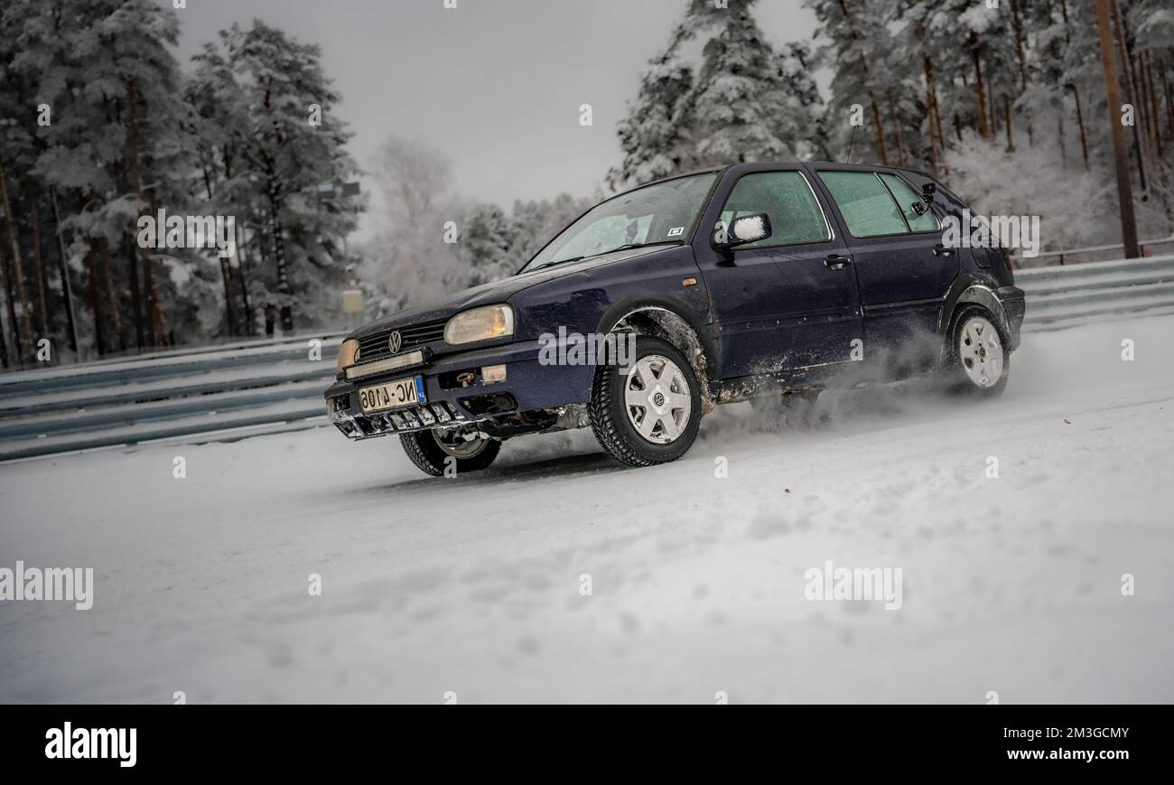 12-12-2022 Riga, Latvia  a blue car driving through a snow covered forest area with trees in the background and a fence in the foreground. . Stock Photo