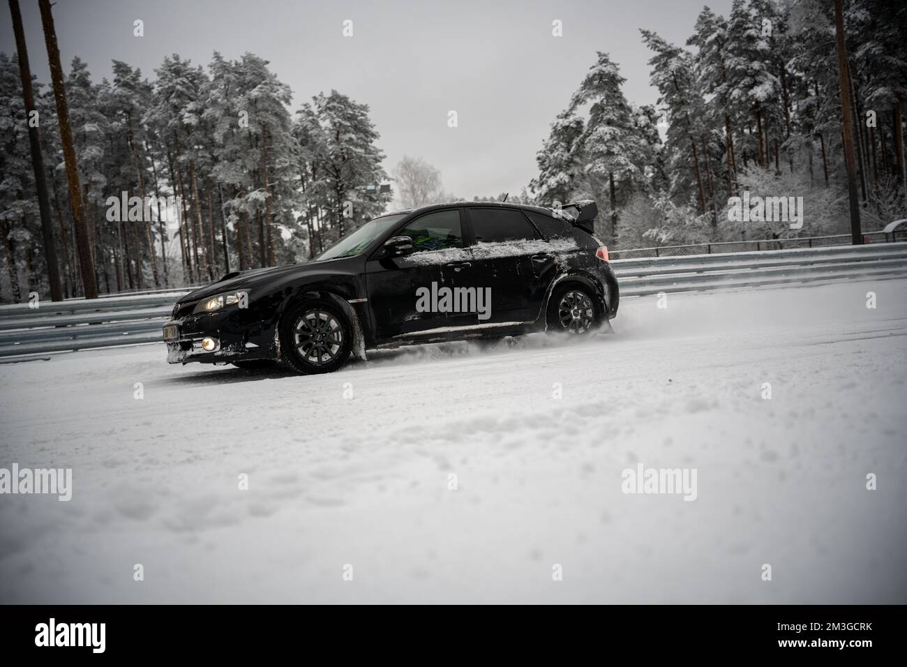 12-12-2022 Riga, Latvia  a black car driving down a snow covered road next to a forest of trees and a fence with snow on it. . Stock Photo