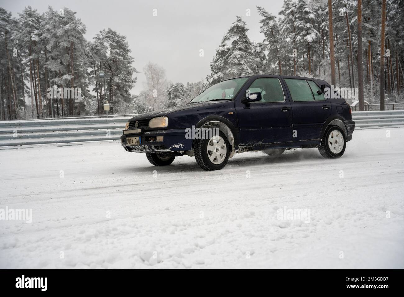 12-12-2022 Riga, Latvia  a black car driving down a snow covered road next to a forest of trees and a fence with snow on it. . Stock Photo
