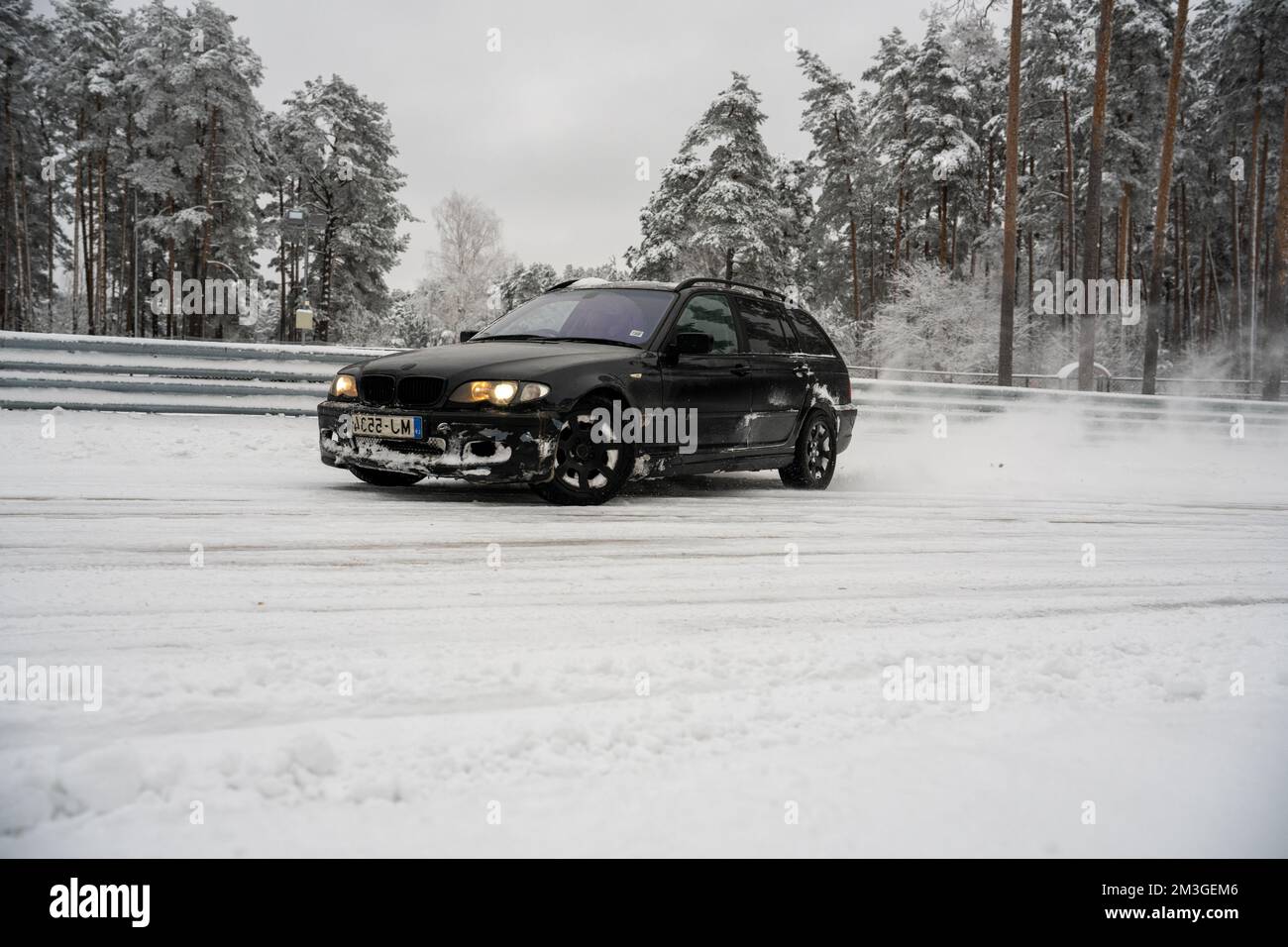 12-12-2022 Riga, Latvia  a black car driving down a snow covered road next to trees and a fence with snow on it and a lot of snow on the ground. . Stock Photo