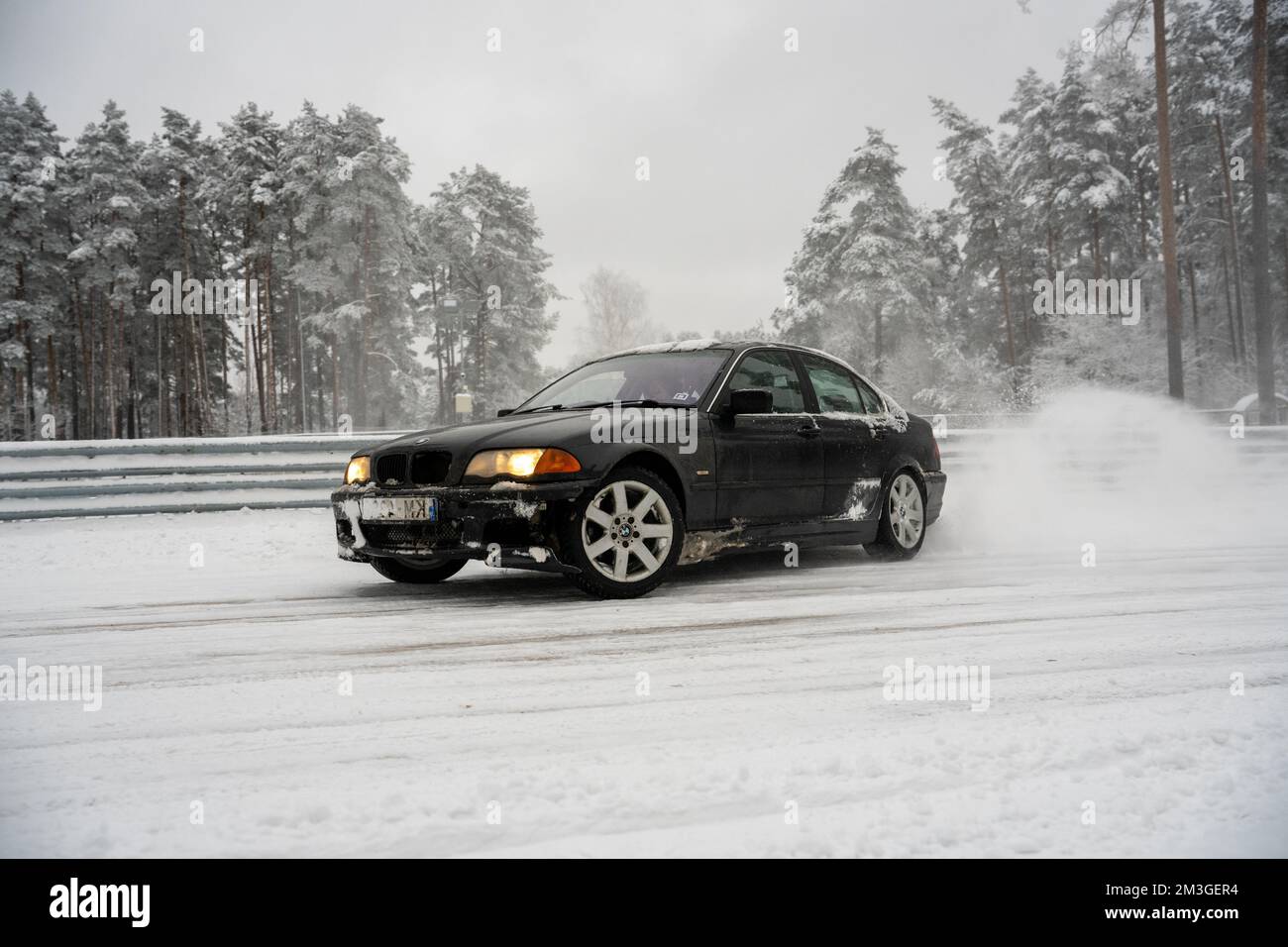 12-12-2022 Riga, Latvia  a black car driving down a snowy road next to a forest of trees and a fence with snow on it. . Stock Photo