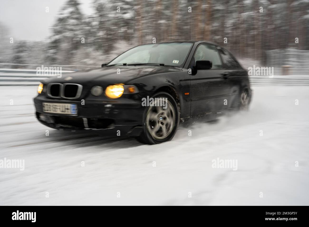 12-12-2022 Riga, Latvia  a black car driving down a snow covered road next to trees and a fence with snow on it and a few cars parked in the snow. . Stock Photo