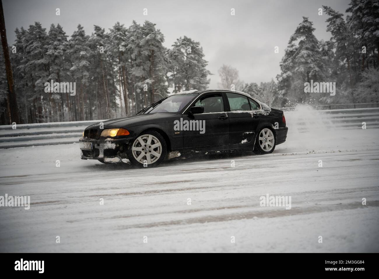 12-12-2022 Riga, Latvia  a black car driving down a snow covered road next to trees and a fence with snow on it and a dusting of snow on the ground. . Stock Photo